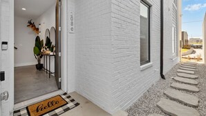 Fachada de ladrillo blanco con un cartel de número de casa, una puerta que da a un interior con una planta y una decoración de cabeza de ciervo, y un camino de piedra afuera.