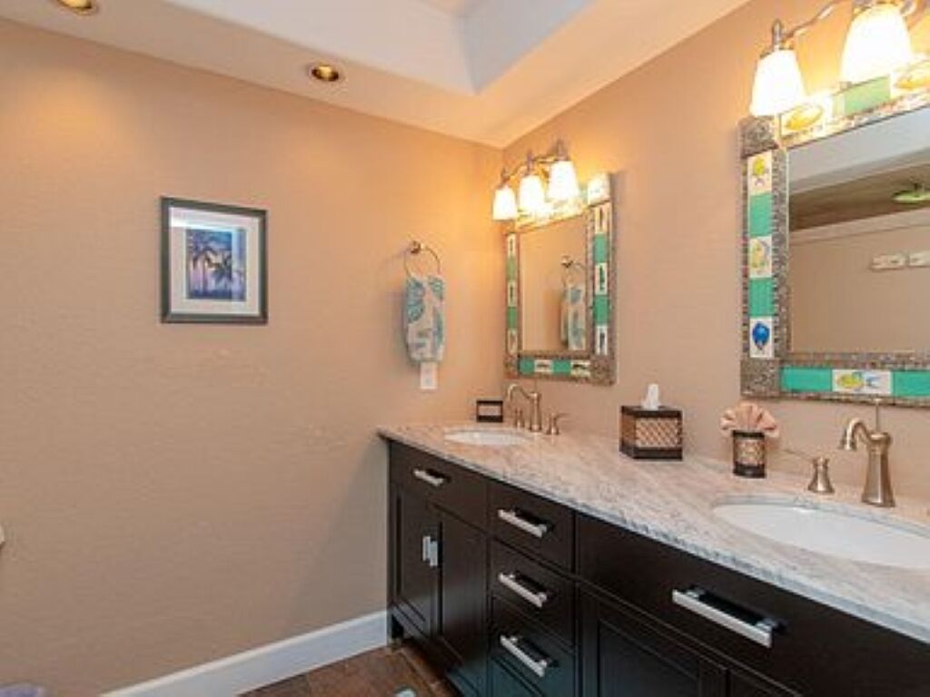A bathroom with a double sink vanity, a large mirror with lights, and a framed picture on the wall.