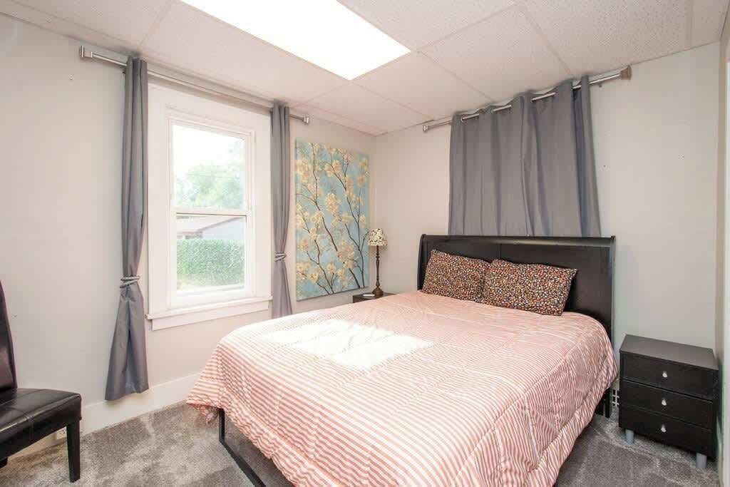 Une chambre à coucher comprenant un lit, une table de chevet, une commode, une fenêtre avec des rideaux et un tableau accroché au mur.