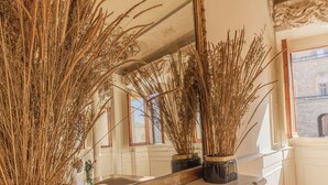 Een kamer met een grote spiegel die hoge, uitgedroogde planten in potten weerspiegelt. Daaronder staat een plank met boeken.