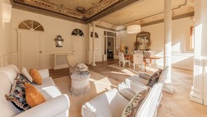 Een ruime woonkamer met een hoge plafond, gedetailleerd plafonddecor en een grote spiegel. Er zijn meerdere zithoeken met witte meubels en een open haard.