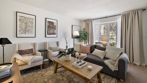 A living room with a sofa, armchairs, a coffee table, and a rug. There are two framed pictures on the wall, a lamp, and a window with curtains. - Closest Stay to Downtown Mt Dora - Renovated - Walk Everywhere (Mount Dora)