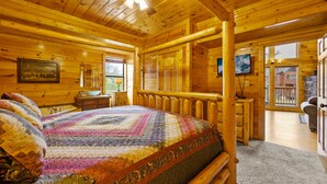 A log cabin bedroom with a large bed, wooden walls, a ceiling fan, and a view of the outside. - 7 min to Dollywood - Hot Tub - Arcades - Sunshine! (Pigeon Forge)