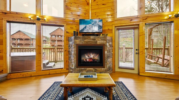 A cozy living room with a stone fireplace, a flat-screen TV mounted above it, and a wooden table in the center. - 7 min to Dollywood - Hot Tub - Arcades - Sunshine! (Pigeon Forge)
