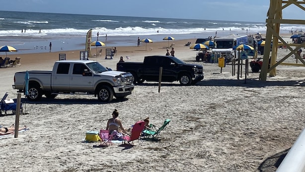 Una playa con una torre de salvavidas, sillas de playa, sombrillas y vehículos.