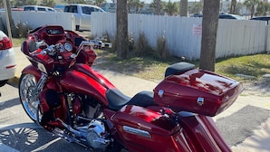 Una motocicleta roja estacionada en un estacionamiento con palmeras y una cerca blanca al fondo.