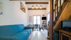 Ein Wohnzimmer mit blauem Sofa, Holzbalken, einem Esstisch mit Stühlen, einem Fernseher und einer Treppe.