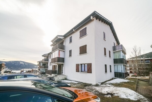 A multi-story residential building with balconies, a parking area with cars, and a mountainous backdrop.