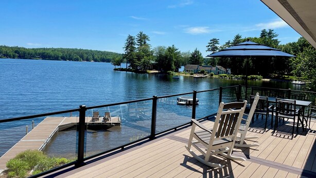 A wooden deck with a view of a lake, a dock, and a small boat. - Lake Winnipesaukee Waterfront Beach & Dock (Moultonborough)