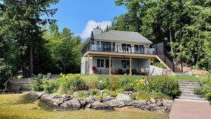 A two-story house with a balcony, surrounded by a pond and various plants. - Lake Winnipesaukee Waterfront Beach & Dock (Moultonborough)