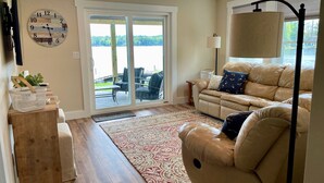 A living room with a sofa, a lamp, a clock, and a view through a glass door to the outdoors. - Lake Winnipesaukee Waterfront Beach & Dock (Moultonborough)