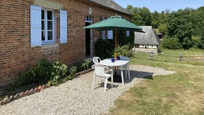 Une maison en briques avec une terrasse en gravier, une table avec des chaises et un parasol vert.