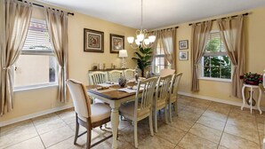 A dining room with a wooden table, beige chairs, and a chandelier. There are two windows with beige curtains, and a plant on the windowsill. - Murano Mews (Kissimmee)
