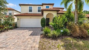 A two-story house with a garage, a brick driveway, and a well-maintained garden with various plants and a palm tree. - Murano Mews (Kissimmee)