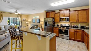 A compact kitchen with granite countertops, wooden cabinets, a stainless steel refrigerator, a microwave, and an oven. - Murano Mews (Kissimmee)