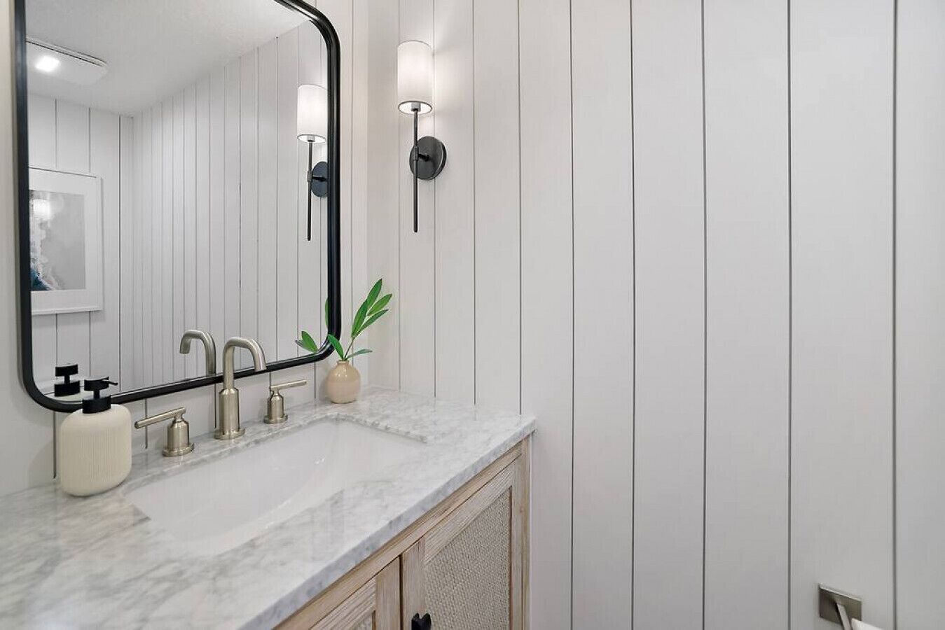 Un baño con tocador blanco, un espejo grande, encimera de mármol, una planta y un aplique de pared.