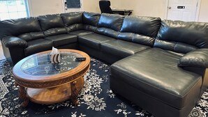 Una sala de estar con un sofá seccional de cuero negro, una mesa de centro de vidrio y un tapete estampado.