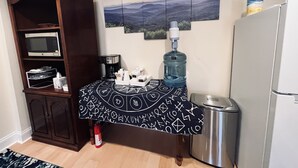 Cocina equipada con microondas, cafetera, dispensador de agua y un cubo de basura de acero inoxidable. Se observa un mueble con un microondas encima y un refrigerador a la derecha.
