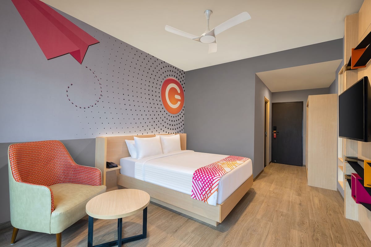 A modern hotel room with a bed, a chair, a small table, a ceiling fan, and a TV.