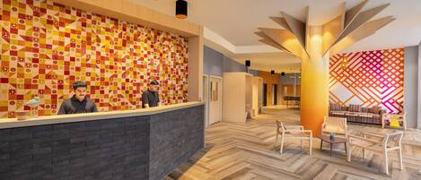 Zona de recepción de hotel con pared estampada, mostrador de recepción y área de estar con una lámpara moderna.