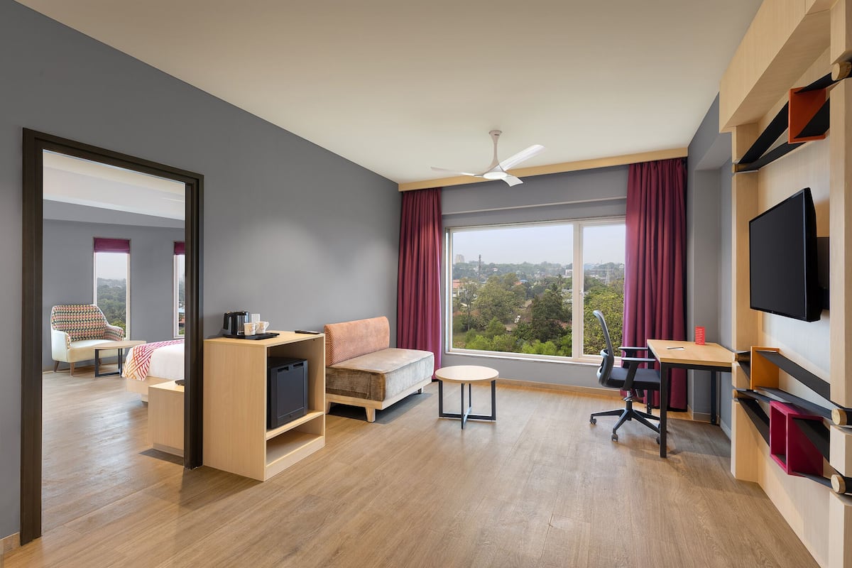 A modern hotel room with a sofa, a small table, a TV, and a large window with a city view.