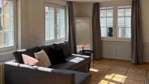 A modern living room with a grey sofa, a small table with a vase, and large windows with curtains. - Hotel Untertor by Maier Self-Check-in (Altstaetten)