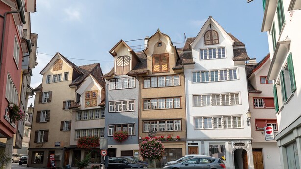A row of multi-story buildings with a central roundabout, cars parked along the street, and flower boxes on the windowsills. - Hotel Untertor by Maier Self-Check-in (Altstaetten)