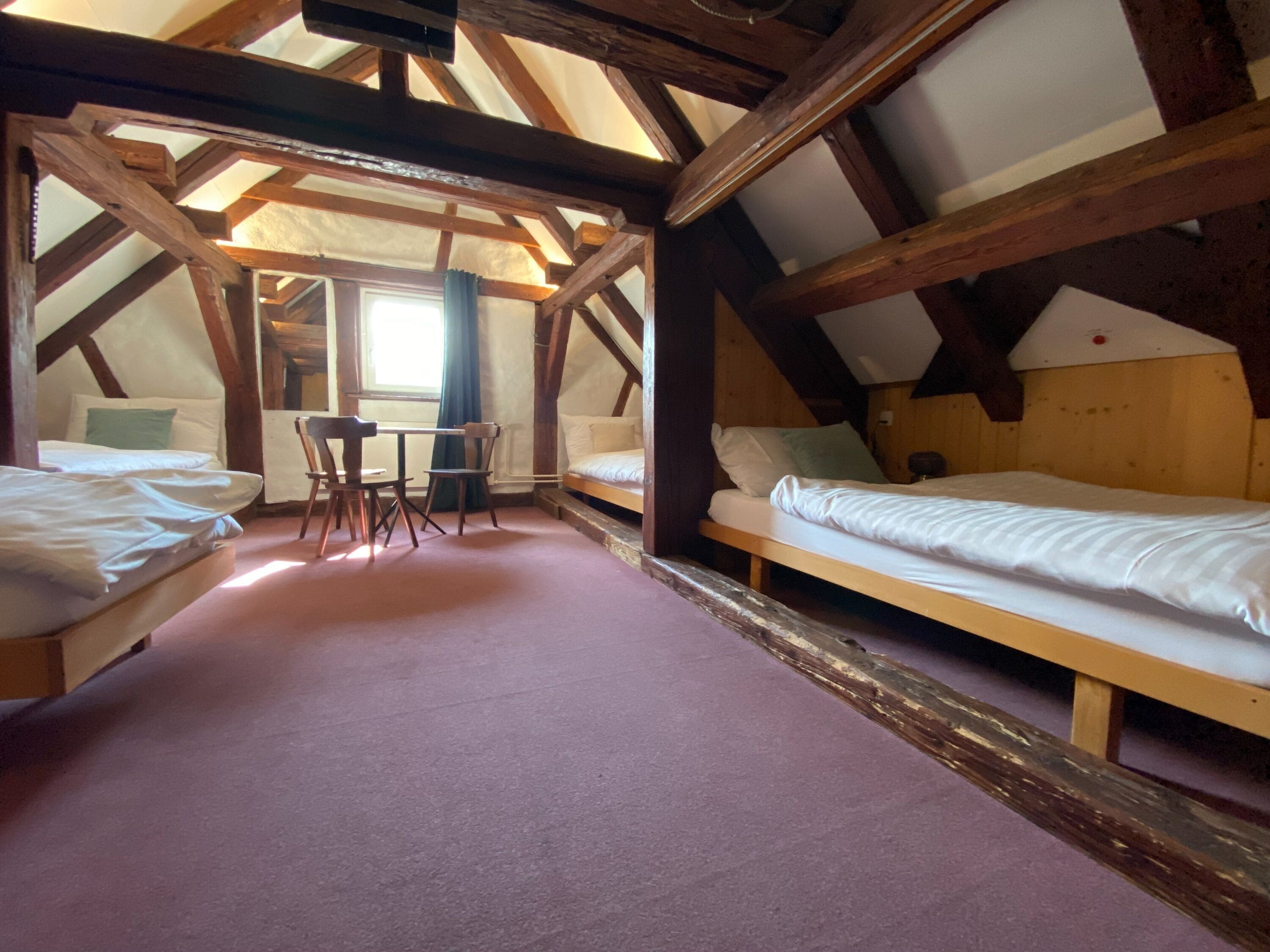 Habitación con dos camas, una mesa y sillas. Cuenta con vigas de madera y techo inclinado.