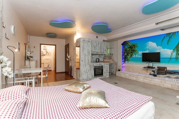 A bedroom with a bed, pillows, a TV, a kitchen area, and a mural of a beach scene.