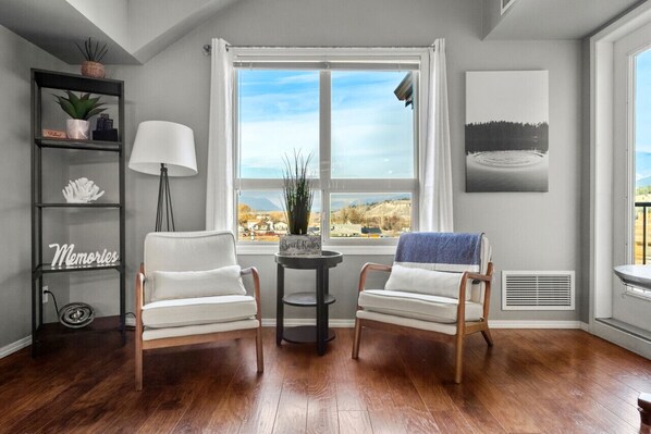 A room with two white armchairs, a small table, a lamp, a shelf with decorative items, and a large window offering a view of a landscape.