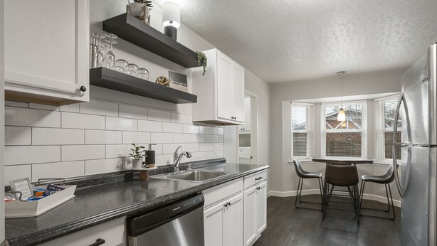 A modern kitchen with white cabinets, a black countertop, stainless steel appliances, and a dining area with a table and chairs. - Charming 3bed/2 bath near Gathering Place, Riverparks and Downtown (Tulsa)