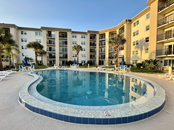A swimming pool with a 'NO DIVING' sign, surrounded by lounge chairs and palm trees, with multi-story buildings in the background.