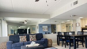 Un salon comprenant un canapé, un pouf et une table à manger avec des chaises. On y trouve également un ventilateur de plafond, un grand miroir et un coin cuisine visible à travers une porte ouverte.