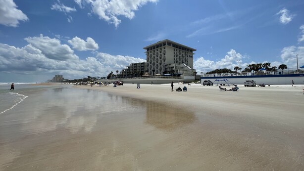 Un hôtel situé en bord de mer, avec un grand bâtiment, une plage de sable et un ciel dégagé.
