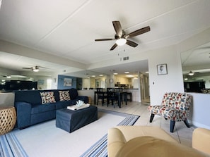 A spacious living area with a sofa, coffee table, and patterned armchair. A ceiling fan is visible, and the room has a large mirror reflecting the space.