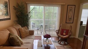 A living room with a beige sofa, a wooden coffee table, a chair with a red cushion, and a large window with blinds. - Reasonable  Rates: Beautiful Condo Close to Beach, Restaurants, Shopping & More! (Rehoboth Beach)