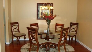 A dining area with a round glass table surrounded by four wooden chairs with cushioned seats. A vase with flowers is placed on the table, and a chandelier hangs above. - Reasonable  Rates: Beautiful Condo Close to Beach, Restaurants, Shopping & More! (Rehoboth Beach)