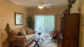 A living room with a beige sofa, a wooden coffee table, a patterned rug, a ceiling fan, and a large window with a view of greenery outside. - Reasonable  Rates: Beautiful Condo Close to Beach, Restaurants, Shopping & More! (Rehoboth Beach)
