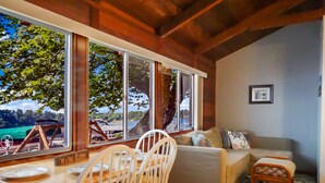 Una habitación acogedora con techo de madera, un sofá, una mesa de comedor con sillas y ventanas amplias que ofrecen vistas al exterior.