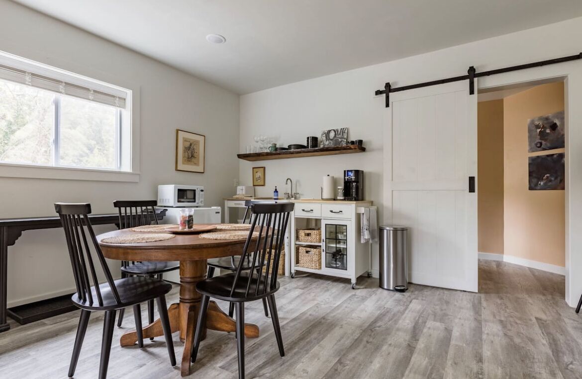 Un coin repas comprenant une table ronde en bois, quatre chaises, un micro-ondes blanc et un petit meuble avec des étagères au-dessus.