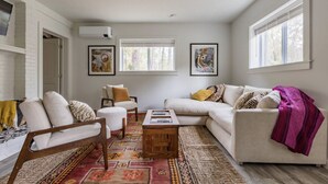 Ein Wohnzimmer mit einem weißen Sofa, einem Holztisch und einem gemusterten Teppich. Zwei Fenster mit Jalousien und Kunstwerken an den Wänden.
