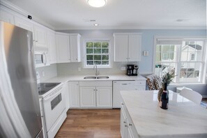 A modern kitchen with white cabinetry, a stainless steel refrigerator, a microwave, and a coffee maker. There is a window providing natural light and a view of the outside. - Spectacular Beach House just 1 min. walk to beach w/ Private Salt Pool & Hot Tub (Surfside Beach)
