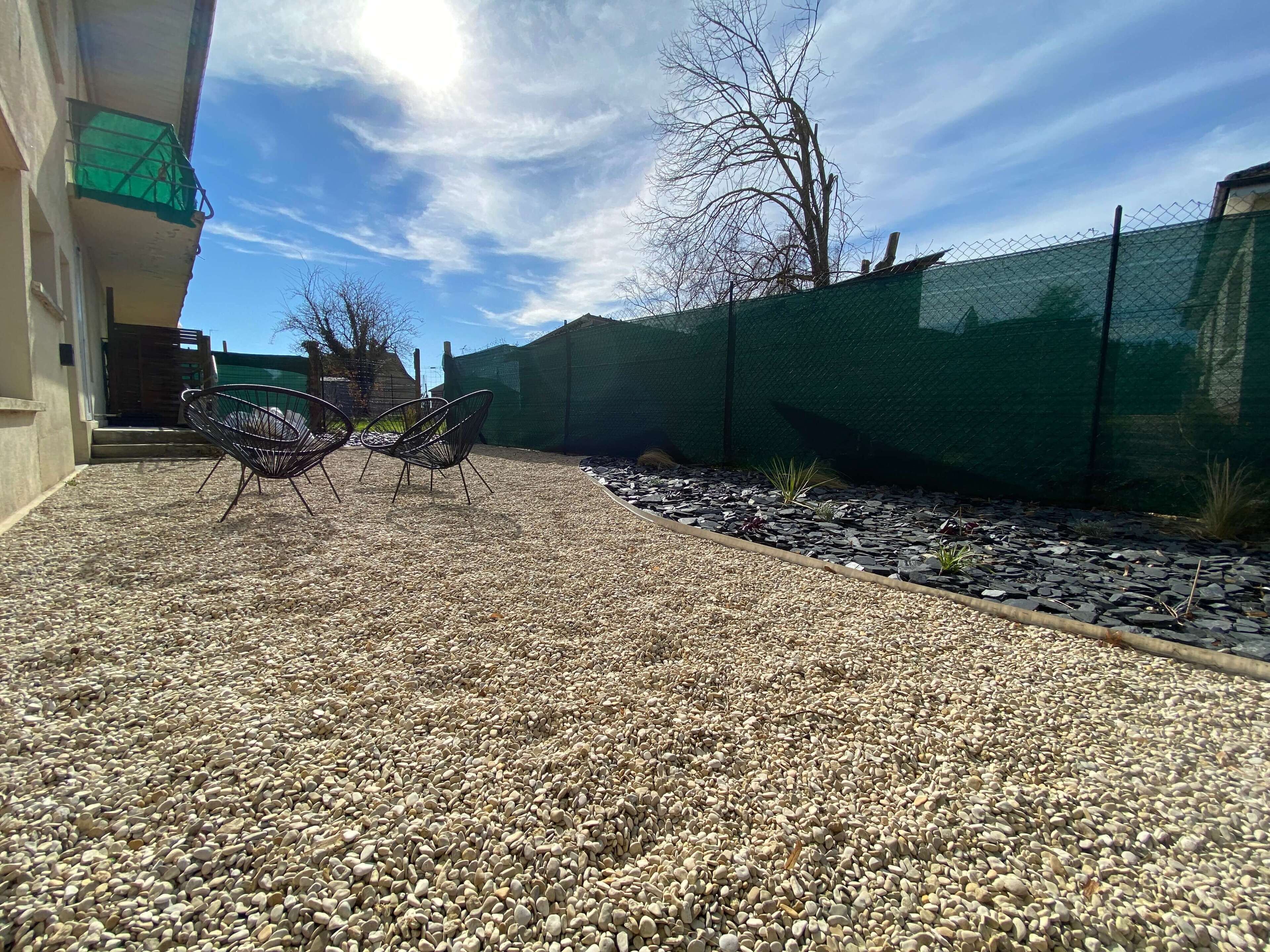 Un espace extérieur aménagé en terrasse, avec un sol en gravier, deux chaises en métal et une clôture verte.