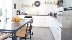 A modern kitchen with white cabinets, a glass dining table, wicker chairs, and a variety of kitchen appliances. - Detached house + private pool located 5 mn from Avignon and 15 mn from St Rémy (Les Angles)