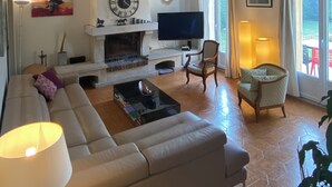 A living room with a sofa, a coffee table, a fireplace, a television, and a large window with curtains. - Detached house + private pool located 5 mn from Avignon and 15 mn from St Rémy (Les Angles)