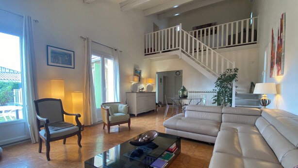 A spacious living room with a large sofa, a glass coffee table, and a dining area with a table and chairs. A staircase leads to an upper level. - Detached house + private pool located 5 mn from Avignon and 15 mn from St Rémy (Les Angles)