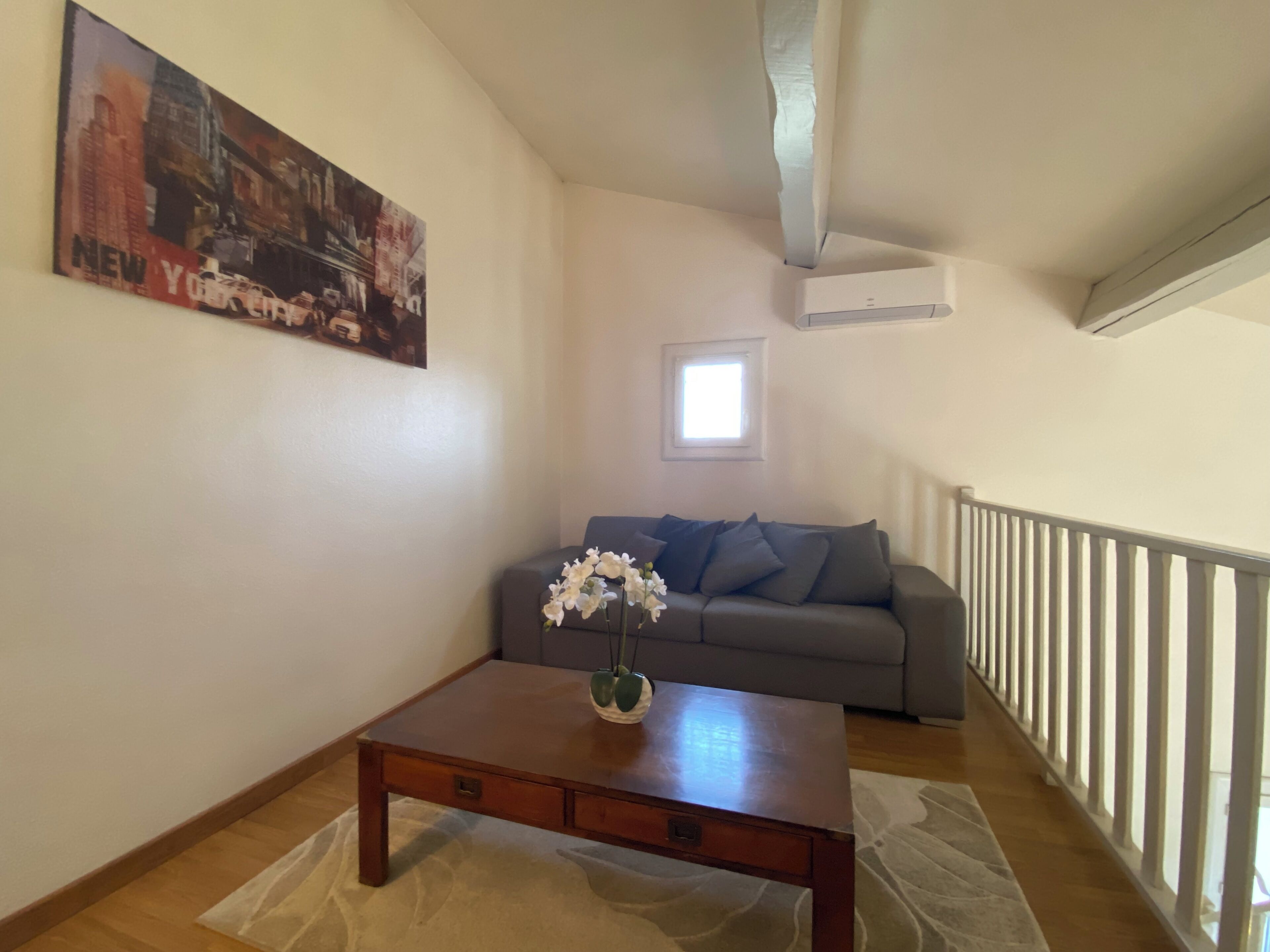 Una sala de estar con una mesa de centro de madera, un sofá gris, un tapete blanco y una pintura de un paisaje urbano en la pared.