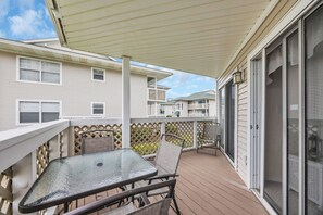 A covered outdoor patio with a glass table and chairs, a wooden deck, and a view of neighboring buildings. - Shorehaven Lakefront & Steps to Beach w Pool (North Myrtle Beach)