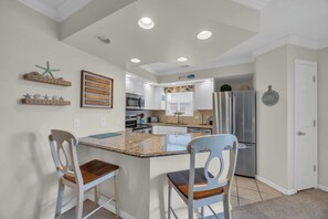 A kitchen with white cabinets, a granite countertop, and a stainless steel refrigerator. There are two bar stools and a small island with a sink. - Shorehaven Lakefront & Steps to Beach w Pool (North Myrtle Beach)