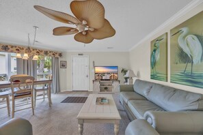 A living room with a sofa, coffee table, dining table, chairs, ceiling fan, artwork, and a television. - Shorehaven Lakefront & Steps to Beach w Pool (North Myrtle Beach)